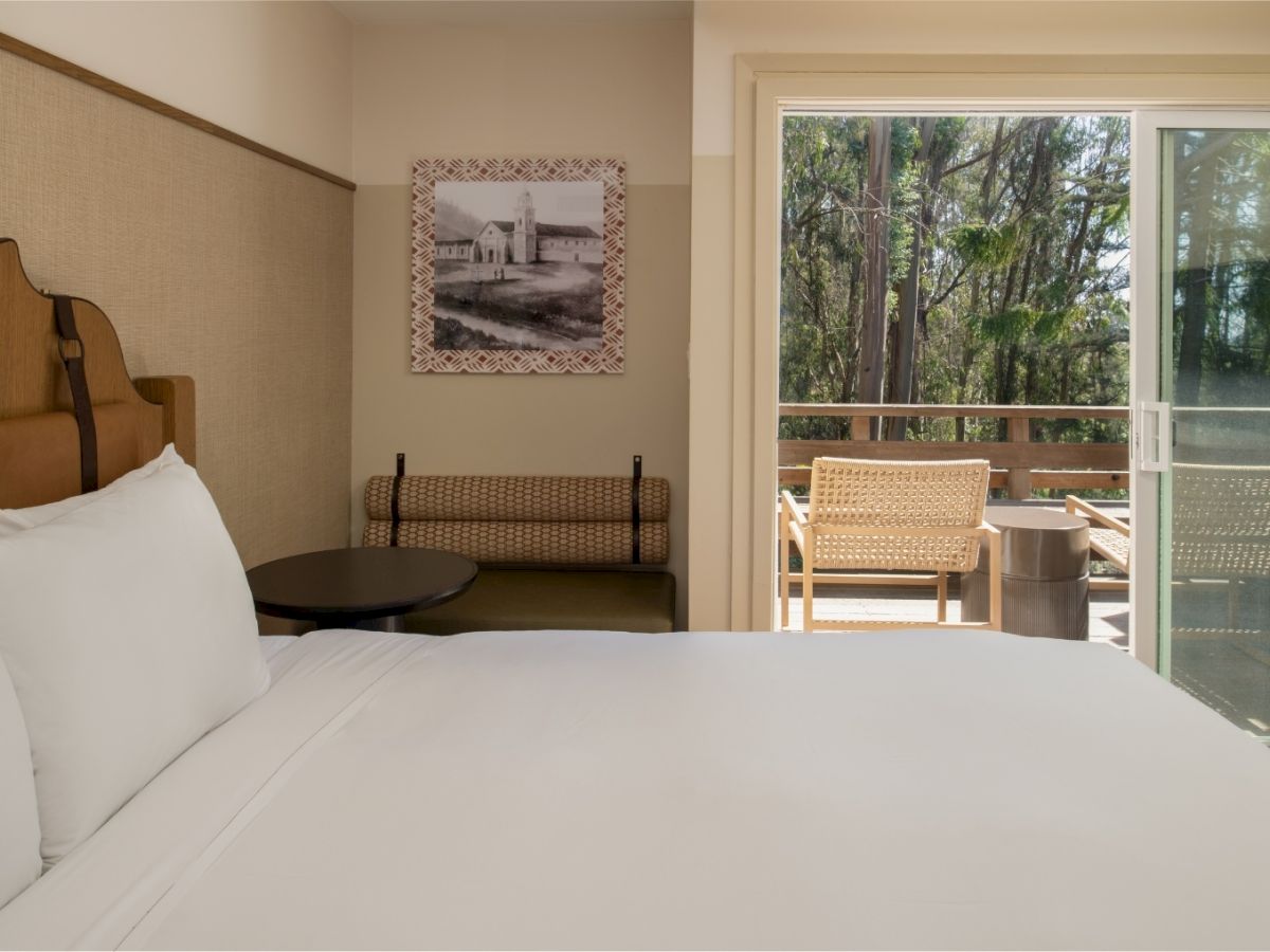 A hotel bedroom with a neatly made bed, wall art, and an open balcony door overlooking green trees.