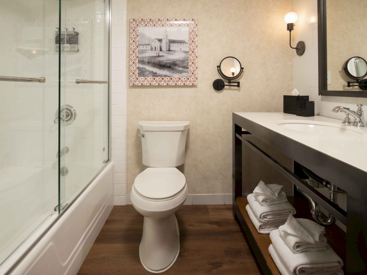 A modern bathroom with a toilet, glass-enclosed shower, and a wide vanity with open shelf holding folded towels, in neutral tones.