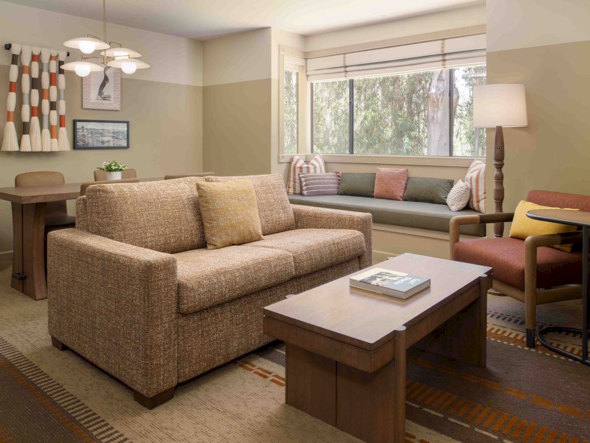 A cozy living room with a beige sofa, a wooden coffee table, a built-in window seat, and a dining area in the background, plus warm lighting.