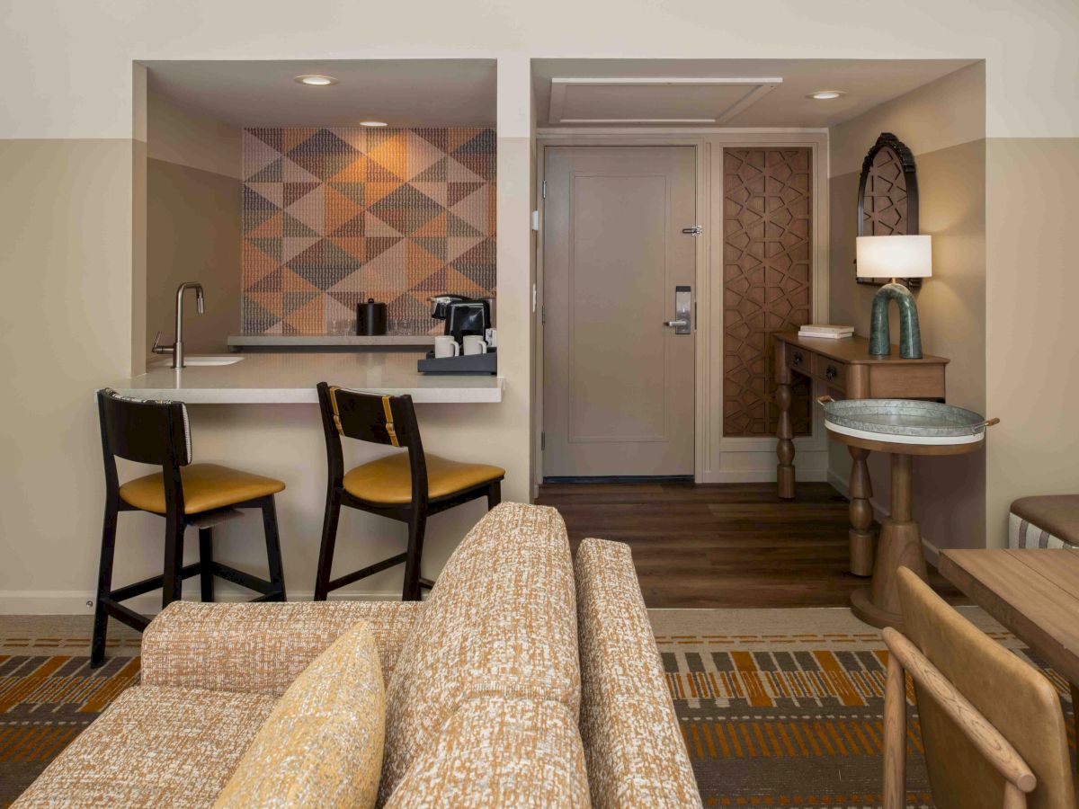 A cozy hotel suite with a kitchenette and breakfast bar, patterned wall accent, sofa, and a small desk area near a doorway.