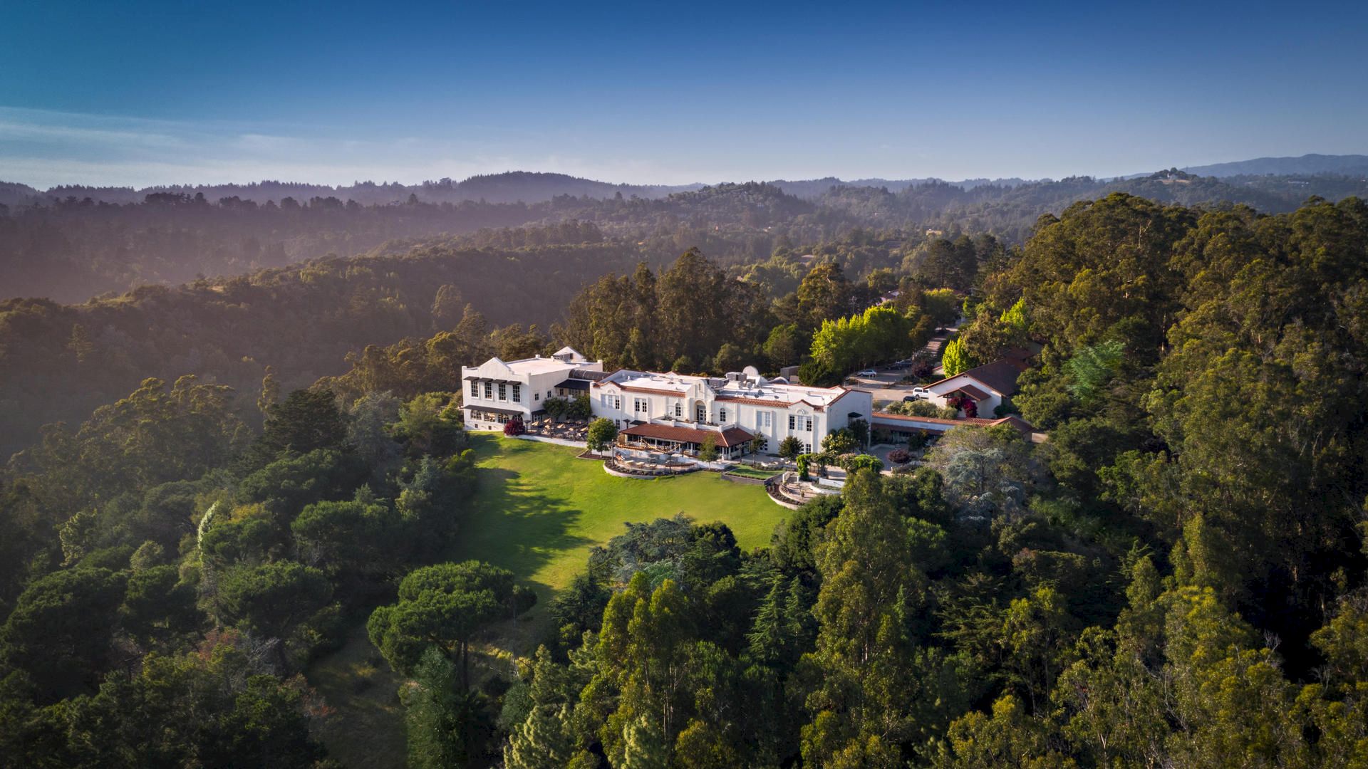 The image shows an aerial view of a large, luxurious mansion surrounded by lush greenery, set within a picturesque landscape, and backed by mountains.